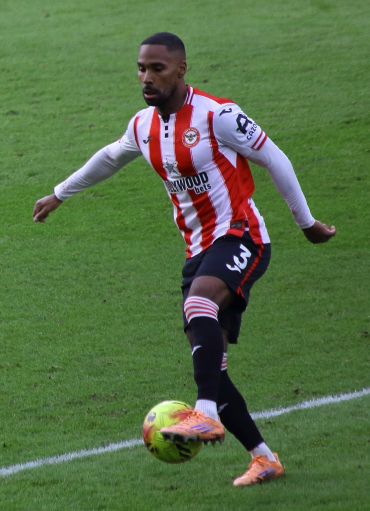 A football player in a red and white striped jersey with the number 3, preparing to kick a ball on a green pitch.