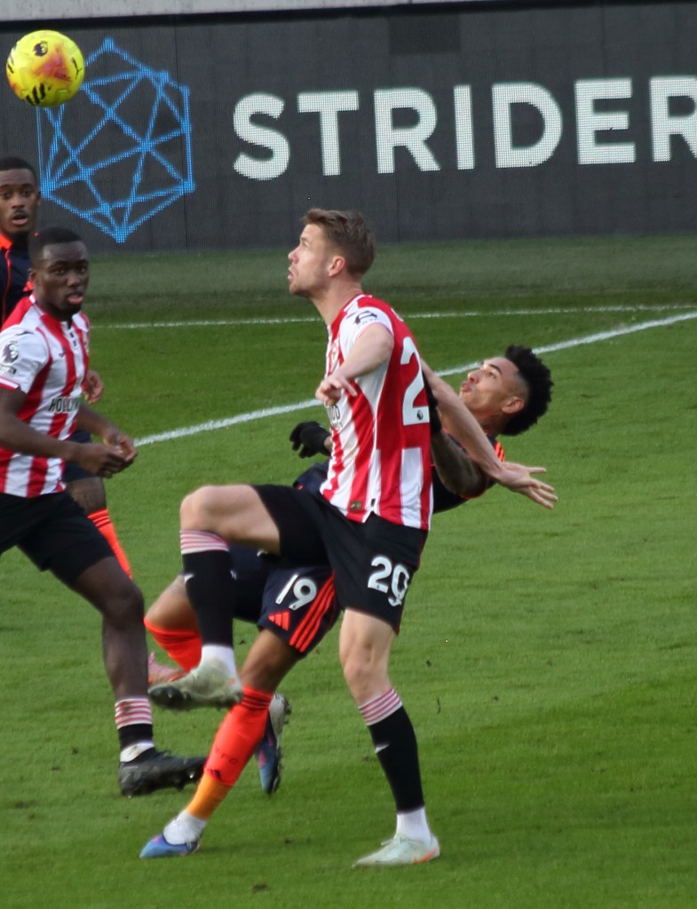 A football match scene showing players from two teams in action, with one player attempting a kick while another player is leaping to head the ball. The background features a scoreboard with the text 'STRIDER'.