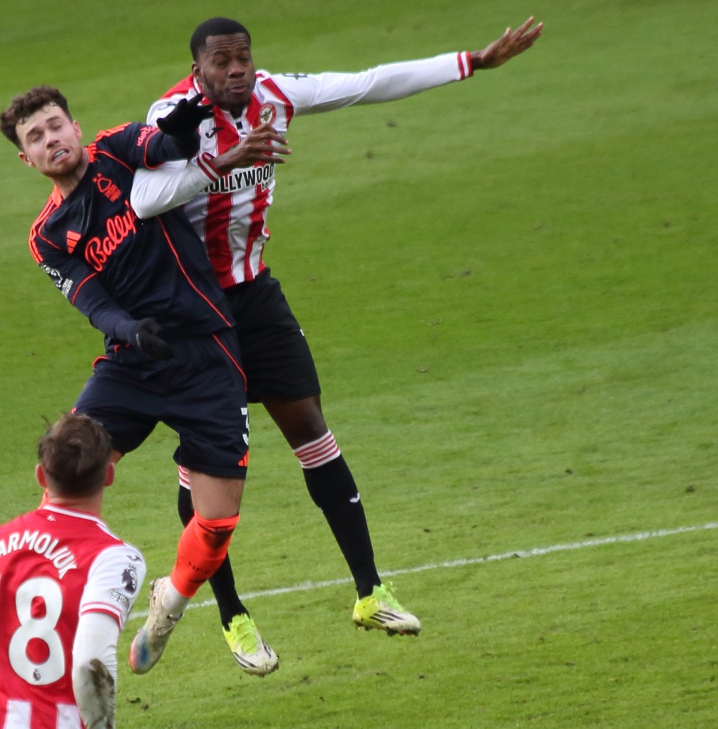 A soccer match scene showing two players grappling for possession of the ball. One player, wearing a dark jersey, is being held by the other player in red and white stripes. Another player is visible in the background.
