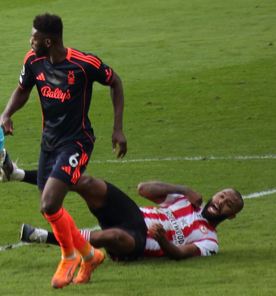 A football match scene showing a player from Nottingham Forest in a black kit with orange accents, evading a fallen player from another team, who is on the ground grimacing in pain.