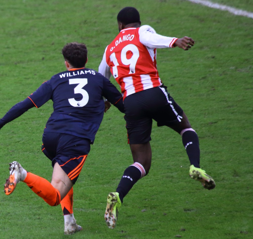 A football match scene showing two players in action, with one in a blue kit labeled 'WILLIAMS' and the other in a red and white striped kit labeled 'DANGO'. The player in blue appears to be lunging towards the player in red and white.