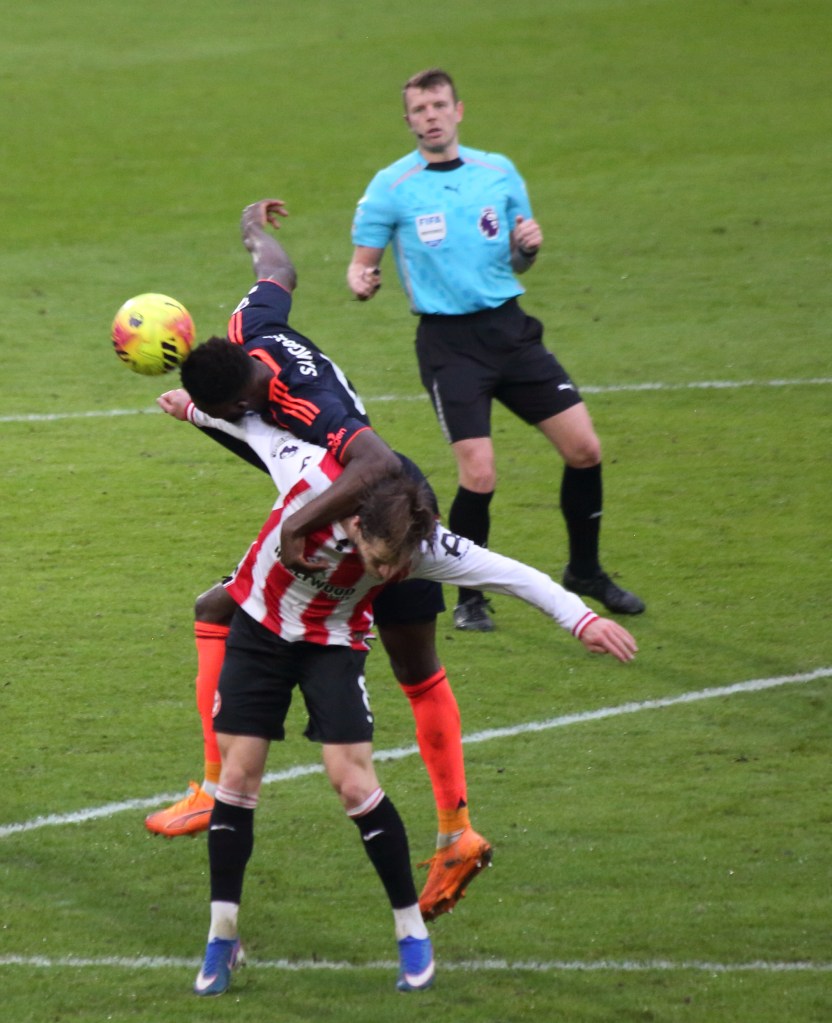 A soccer match scene depicting two players in a physical contest for the ball, with one player heading the ball and another in a defensive position. An official is observing the play in the background.