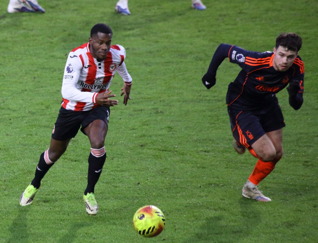 Two soccer players compete for the ball on the field during a match, with one player in a red and white striped jersey and the other in a dark jersey.